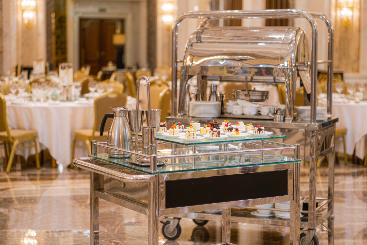 Stainless steel and glass hotel banquet service trolley displaying plated desserts and a coffee service in front of a three tier stainless steel catering trolley in a ballroom