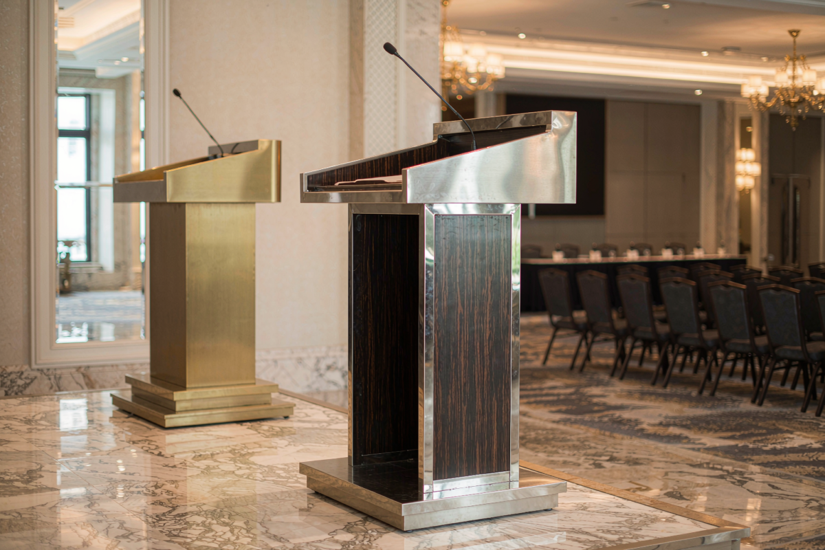 Two hotel podiums on a marble conference floor: a brushed gold lectern and a stainless steel lectern with dark wood panels, facing rows of chairs in a ballroom